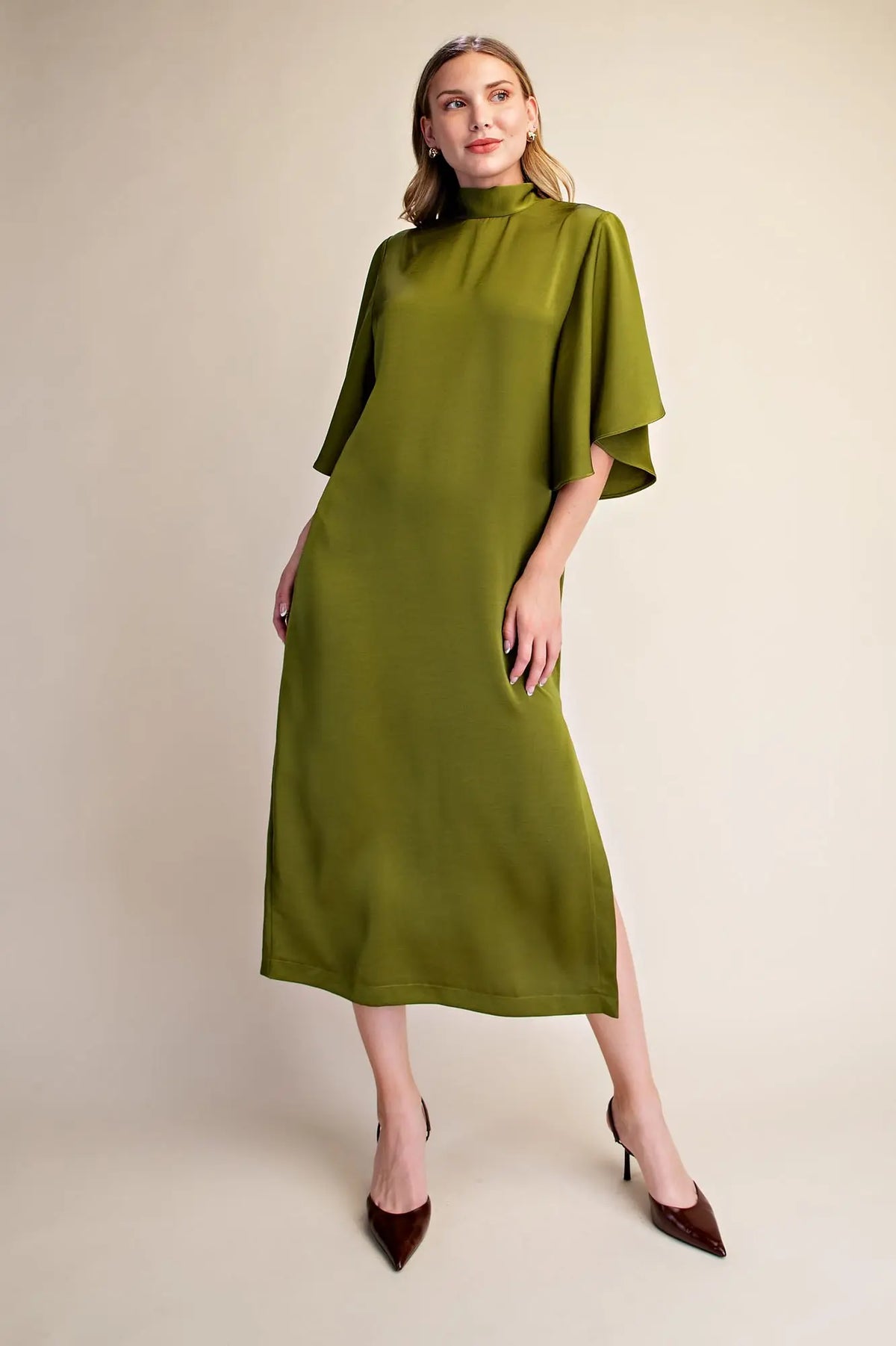 A woman models a high-neck, olive green midi dress with wide, draped half sleeves and a side slit, paired with dark brown pointed-toe heels against a plain beige studio background.