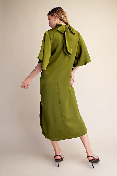 A woman modeling a sage green midi dress with wide flutter sleeves and a large bow detail at the back neckline, shown from a rear three-quarter angle against a plain beige studio background.
