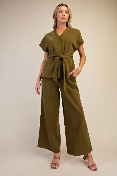 A woman modeling an olive green belted jumpsuit with short sleeves and wide-leg pants, standing against a plain beige studio background.