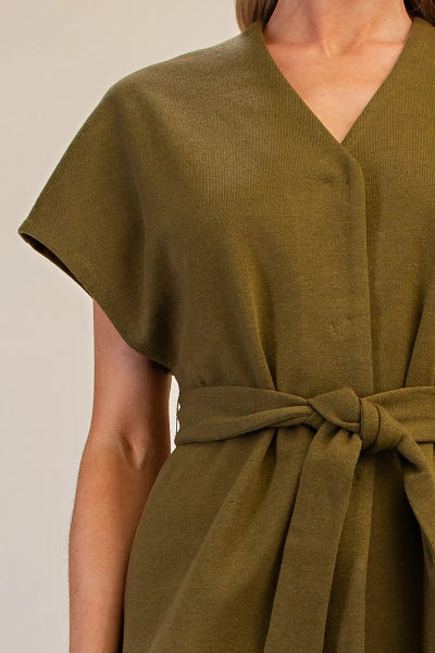 Close-up of a model wearing an olive green belted wrap dress with short sleeves and a V-neckline against a plain light beige background.
