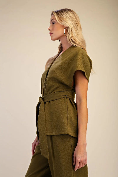 A blonde woman in profile wearing an olive green belted wrap-style top and matching wide-leg trousers, photographed against a plain warm beige studio background.