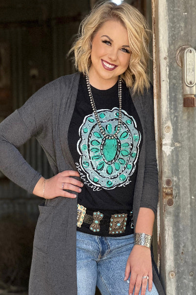 Woman wearing a black t-shirt with a green design, gray cardigan, and blue jeans in front of a rustic metal door.