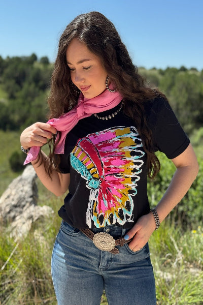 Woman wearing a black t-shirt with a colorful headdress design, pink scarf, and blue jeans outdoors.