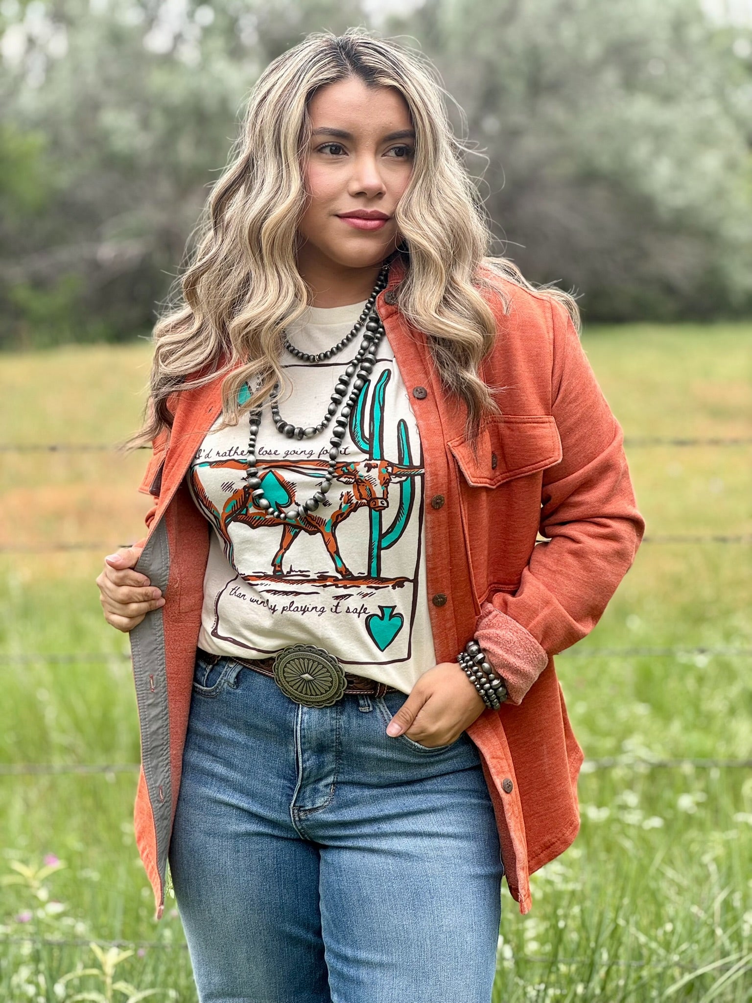 Woman wearing an orange jacket, graphic t-shirt, and blue jeans in a field.