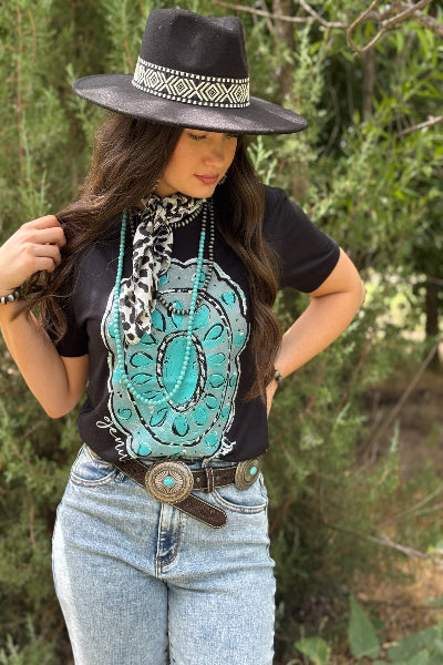 Woman wearing a black hat, black shirt with turquoise design, and light blue jeans outdoors.
