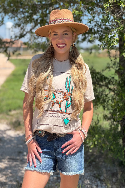 Woman wearing a beige t-shirt with a graphic design, denim shorts, and a wide-brimmed hat outdoors.