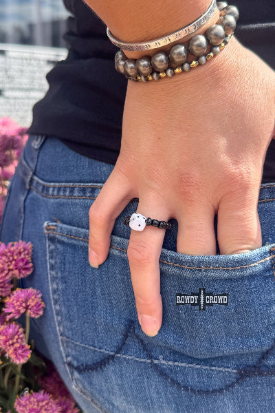 A close-up lifestyle shot of a person's hand in a denim pocket, wearing a black band ring with a white cat face. The person is also wearing a silver beaded bracelet and a black top, with pink flowers visible on the left side available at Cowtown Bling N Things.