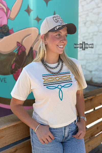 A woman wearing a cream-colored graphic t-shirt with a teal and gold geometric design, paired with a beige and brown baseball cap and blue jeans, posing outdoors in front of a colorful mural available at Cowtown Bling N Things.