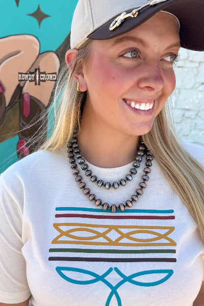 A woman wearing a white graphic t-shirt with a colorful geometric pattern, a multi-strand beaded necklace, and a baseball cap, standing in front of a colorful mural available at Cowtown Bling N Things.