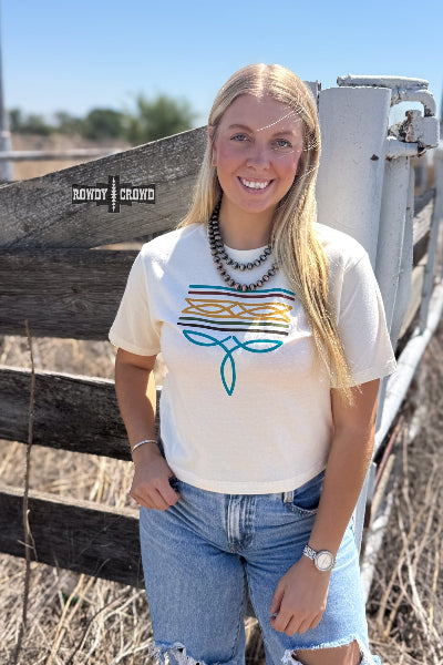 A smiling blonde woman stands outdoors in front of a weathered wooden fence, wearing a cream graphic t-shirt with a blue and orange geometric design, light-wash jeans, and layered beaded necklaces. The brand name 'ROWDY CROWD' is visible as a watermark on the left side of the image available at Cowtown Bling N Things.