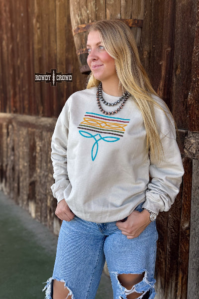 A woman wearing a cream-colored crewneck sweatshirt with a colorful striped and bow graphic on the front, paired with ripped blue jeans, standing against a rustic wooden wall. The brand name 'Rowdy Crowd' is visible as a watermark on the left side available at Cowtown Bling N Things.