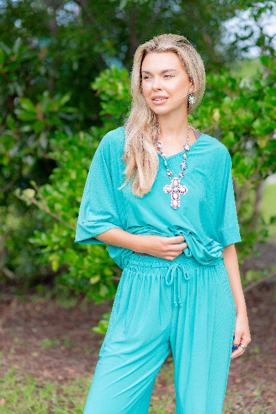 Woman wearing a turquoise jumpsuit with a necklace, standing outdoors with greenery in the background