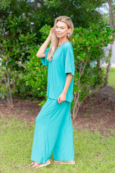 Woman wearing a teal outfit standing in a natural setting with greenery.