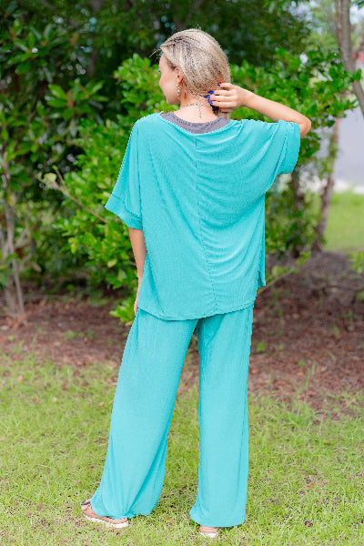 Person wearing a teal outfit standing outdoors with greenery in the background