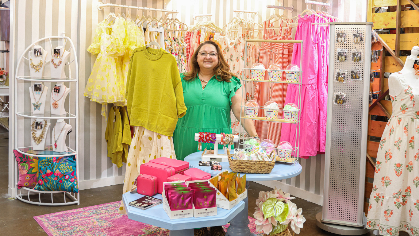 A woman stands in a boutique interior surrounded by clothing racks, mannequins, and a small display table with colorful accessories. The scene includes striped display boards, floral garments, jewelry stands, and bright retail decor.