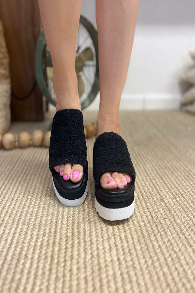 Black open-toe sandals with white soles worn indoors on a carpeted floor.