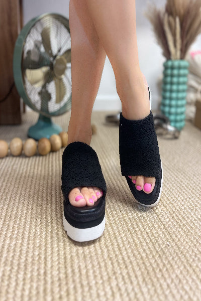 Black open-toe sandals worn indoors with a fan and decorative items in the background.