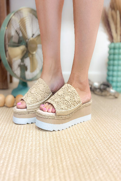 Beige sandals with white soles worn by a person on a textured floor.
