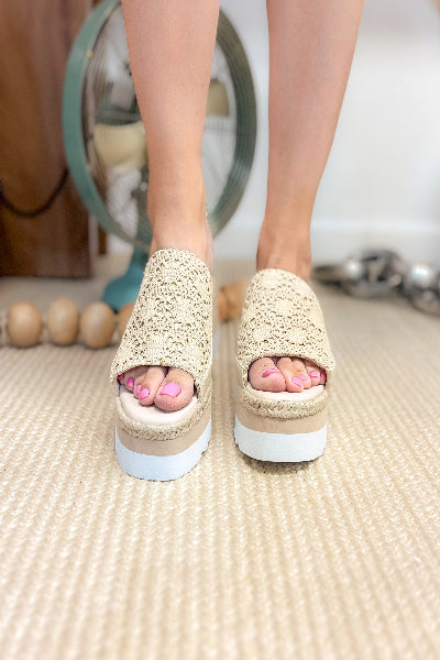 Beige sandals with white soles worn by a person on a textured floor.