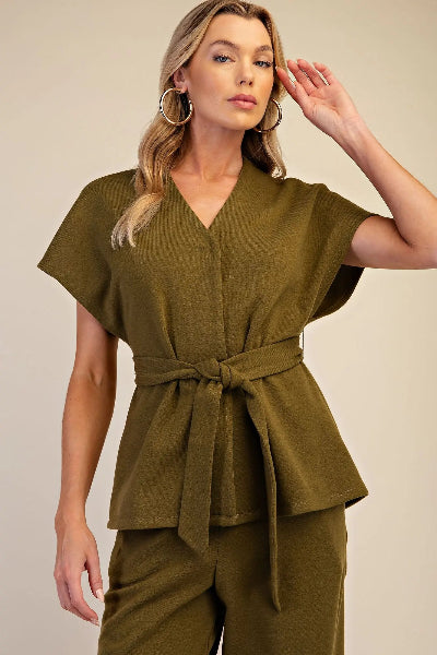 A woman modeling a short-sleeve olive green belted top and matching pants against a plain beige studio background.