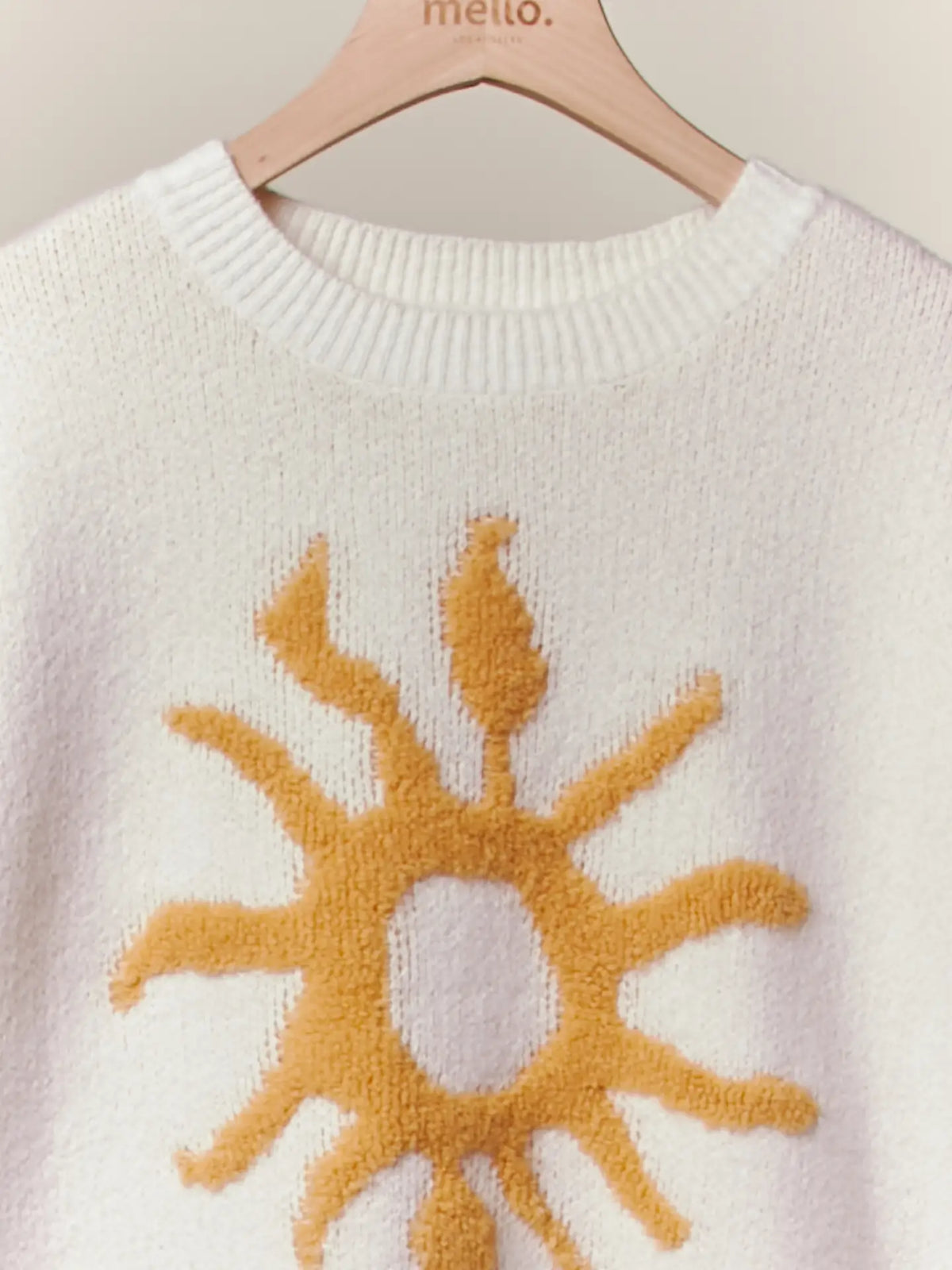 A close-up of a cream-colored knit sweater with a textured mustard yellow sun motif on the front, hanging on a light wood hanger with the brand name 'mello.' visible at the collar.