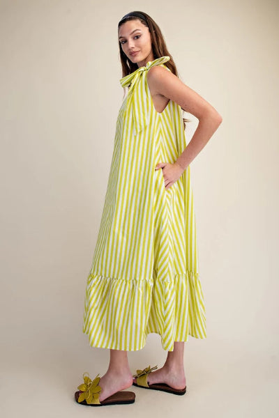 A woman modeling a sleeveless yellow and white striped midi dress with a tie-neck detail and ruffled hem, paired with yellow flower slide sandals, against a plain beige studio background available at Cowtown Bling N Things.