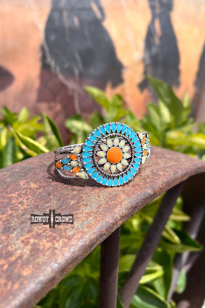 Decorative ring with turquoise and orange stones on a metal surface with greenery in the background