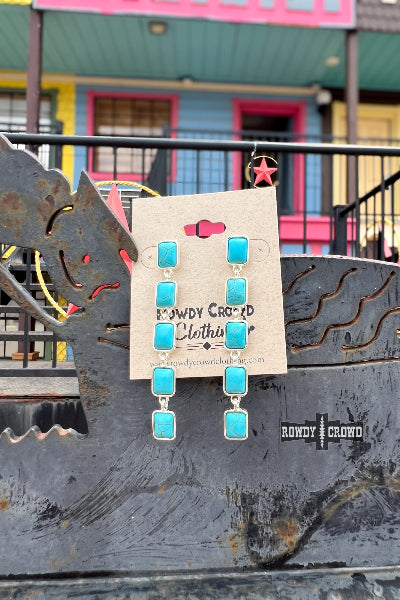 Turquoise earrings with a 'Rowdy Crowd' label on a metal surface with colorful building background