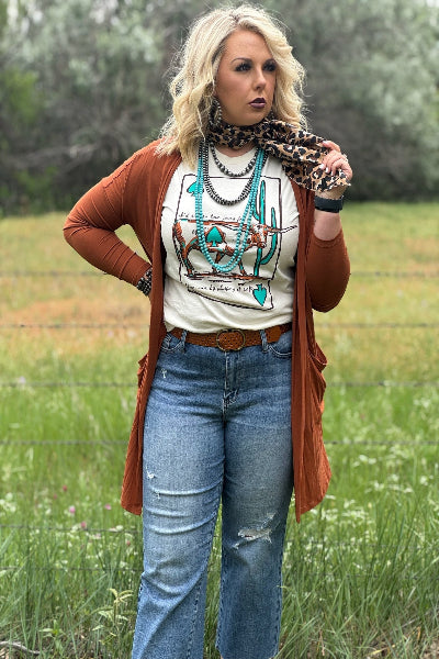 Woman standing in a field wearing a white graphic t-shirt, blue jeans, and a brown cardigan.