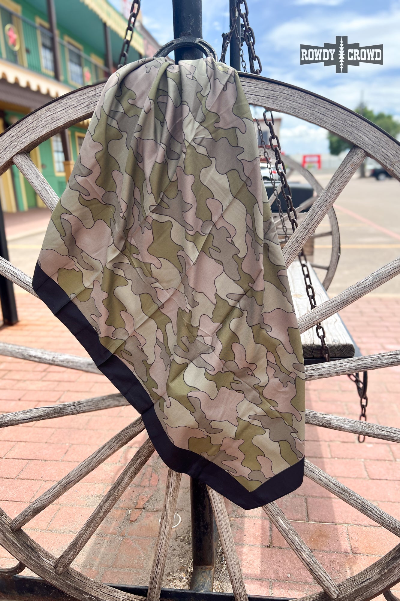 Camouflage-patterned blanket draped over a wooden wagon wheel with a western-style building in the background.