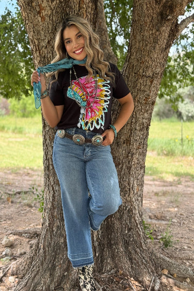 Woman in a colorful top and jeans standing next to a tree in an outdoor setting