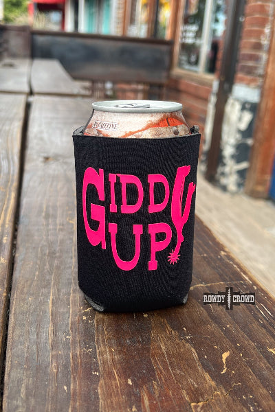A black can cooler sleeve with bright pink 'GIDDY GUP!' lettering, holding a can on a rustic wooden table with a blurred outdoor background available at Cowtown Bling N Things.