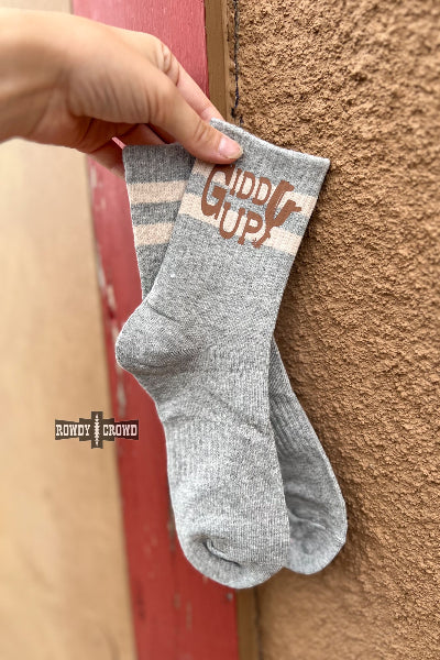 A person's hand holds a pair of heather gray crew socks with white stripes and a brown 'GIDDY UP!' graphic on the cuff, leaning against a textured brown stucco wall next to a red wooden door.