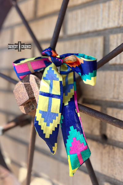 A colorful patterned silk scarf tied in a bow and hanging from a rustic metal wheel, set against a brick wall background. The scarf features a geometric pixelated design in blue, yellow, pink, and green.