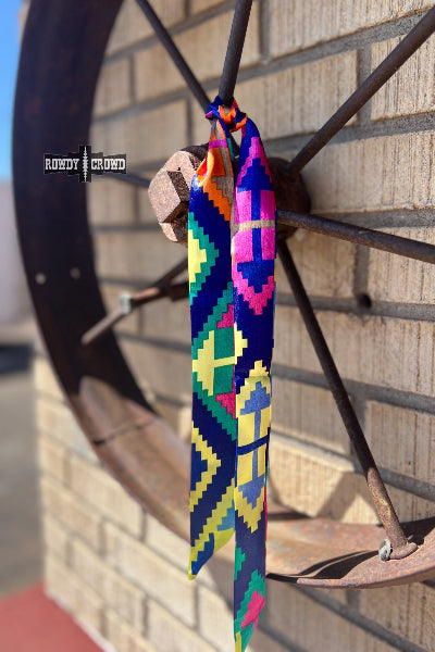 A colorful geometric patterned tie or scarf hanging from a rustic metal wagon wheel against a brick wall. The tie features a vibrant pixelated pattern in blue, pink, yellow, green, and orange available at Cowtown Bling N Things.