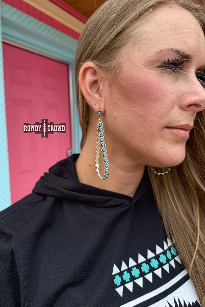 Woman wearing long turquoise earrings with a black hoodie featuring a geometric pattern.