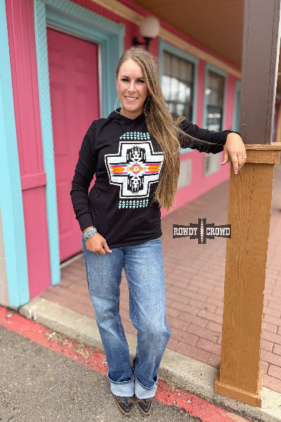 Woman wearing a black hoodie with a graphic design, standing in front of a colorful building.
