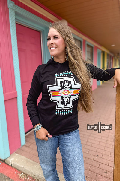Woman wearing a black long-sleeve shirt with a graphic design, standing in front of a colorful building.