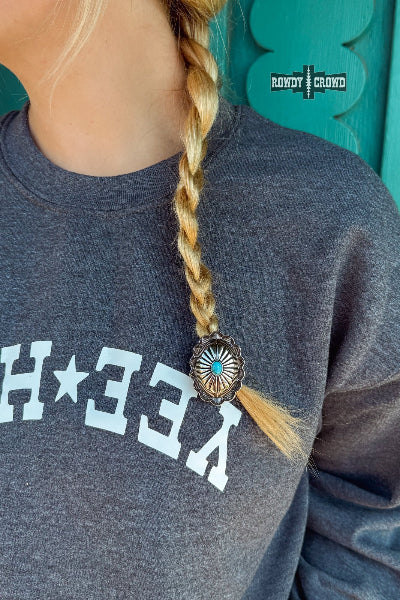 A close-up lifestyle shot of a person wearing a dark heather gray sweatshirt with white graphic lettering and a blonde braid adorned with a western-style concho shell hair clip. The background is a teal wooden door, and a 'Rowdy Crowd' logo is visible on the upper right available at Cowtown Bling N Things.