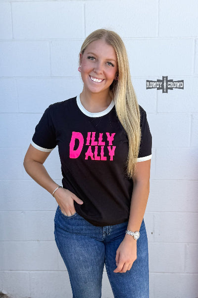 A smiling blonde woman wearing a black ringer t-shirt with white trim and a bright pink 'DILLY ALLY' graphic, paired with blue jeans, standing against a white painted brick wall.