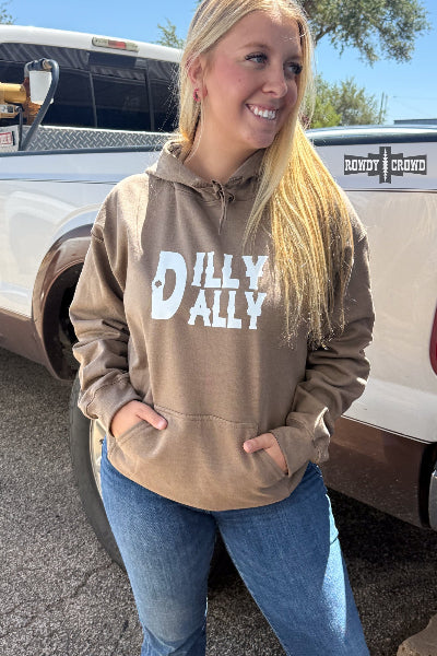 A smiling blonde woman wearing a brown pullover hoodie with 'DILLY DALLY' printed in white across the chest, standing outdoors next to a white pickup truck.
