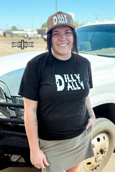 A smiling woman stands outdoors in front of a white pickup truck, wearing a black graphic T-shirt and a brown baseball cap. Both the shirt and cap feature the words 'DILLY ALLY' in large white letters available at Cowtown Bling N Things.