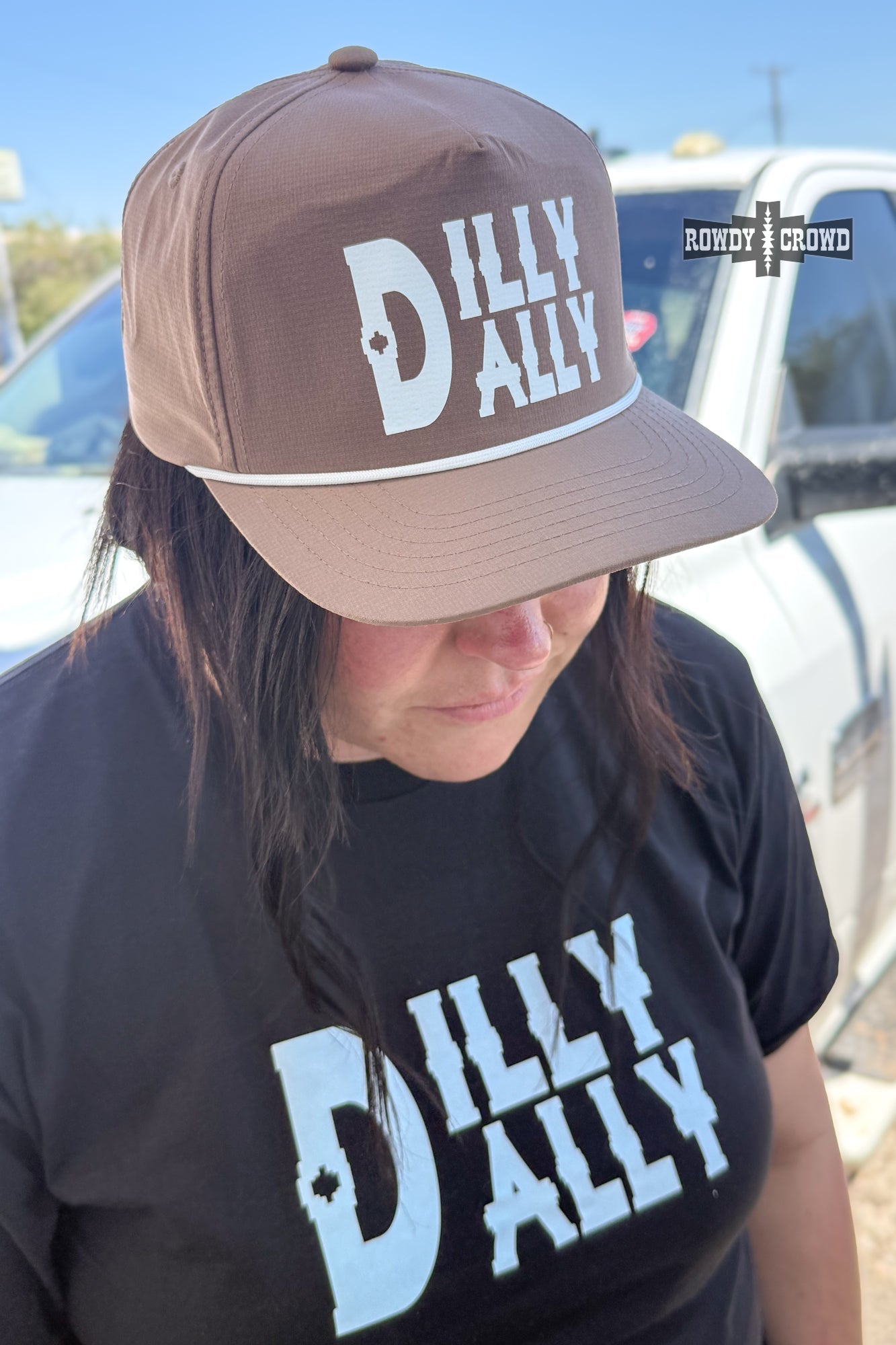 A person outdoors wearing a brown baseball cap with a white 'DILLY DALLY' graphic and a matching black T-shirt with a large light blue 'DILLY DALLY' print. A white pickup truck and blue sky are visible in the background.