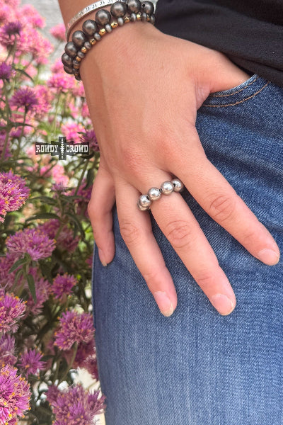 A close-up lifestyle shot of a person's hand wearing a silver ring with three polished round beads, styled with a beaded bracelet and denim jeans against a background of pink flowers available at Cowtown Bling N Things.