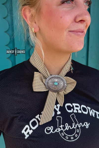 A close-up lifestyle shot of a woman wearing a western-style striped bolo tie with a large silver-toned concho buckle and turquoise center, paired with a black graphic t-shirt available at Cowtown Bling N Things.