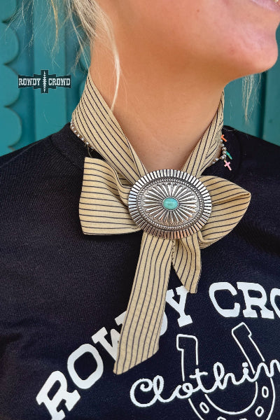 A close-up of a person wearing a beige and black striped neck scarf tied in a large bow with a large silver concho-style brooch featuring a turquoise center. The person is also wearing a black graphic t-shirt with 'Rowdy Crowd Clothing' printed on it, and a turquoise wall is visible in the background available at Cowtown Bling N Things.