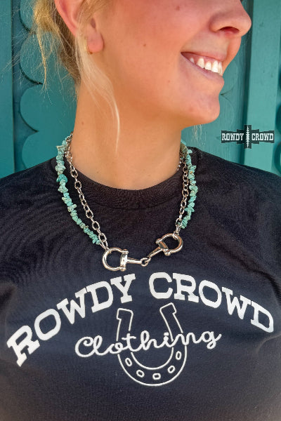 A close-up lifestyle shot of a woman wearing a silver horse-bit style necklace with turquoise beads, layered over a black t-shirt with a white 'ROWDY CROWD' graphic. The background is a teal wooden door available at Rowdy Crowd Clothing.
