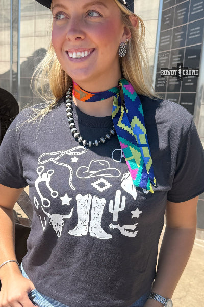 A woman wearing a dark gray graphic t-shirt with a white western-themed print, accessorized with a colorful patterned neck scarf and a beaded necklace available at Cowtown Bling N Things.