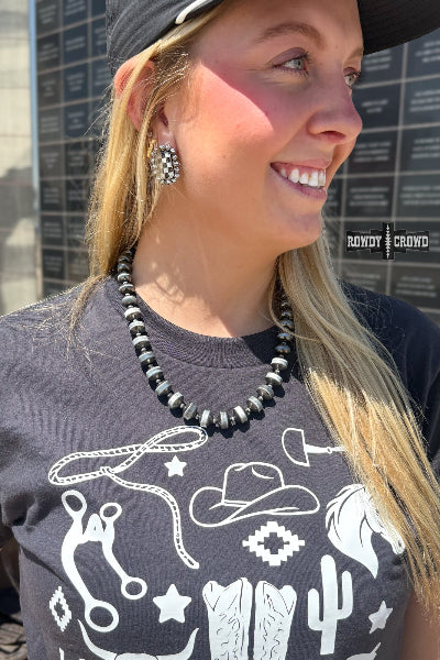 A smiling blonde woman wearing a black beaded necklace, a black graphic t-shirt with white western-themed illustrations, and a black cap, photographed outdoors against a dark stone wall available at Cowtown Bling N Things.