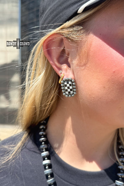 A close-up lifestyle shot of a woman wearing a checkered earring. The earring features a black and white checkerboard pattern surrounded by silver-toned metal and small clear rhinestones available at Cowtown Bling N Things.